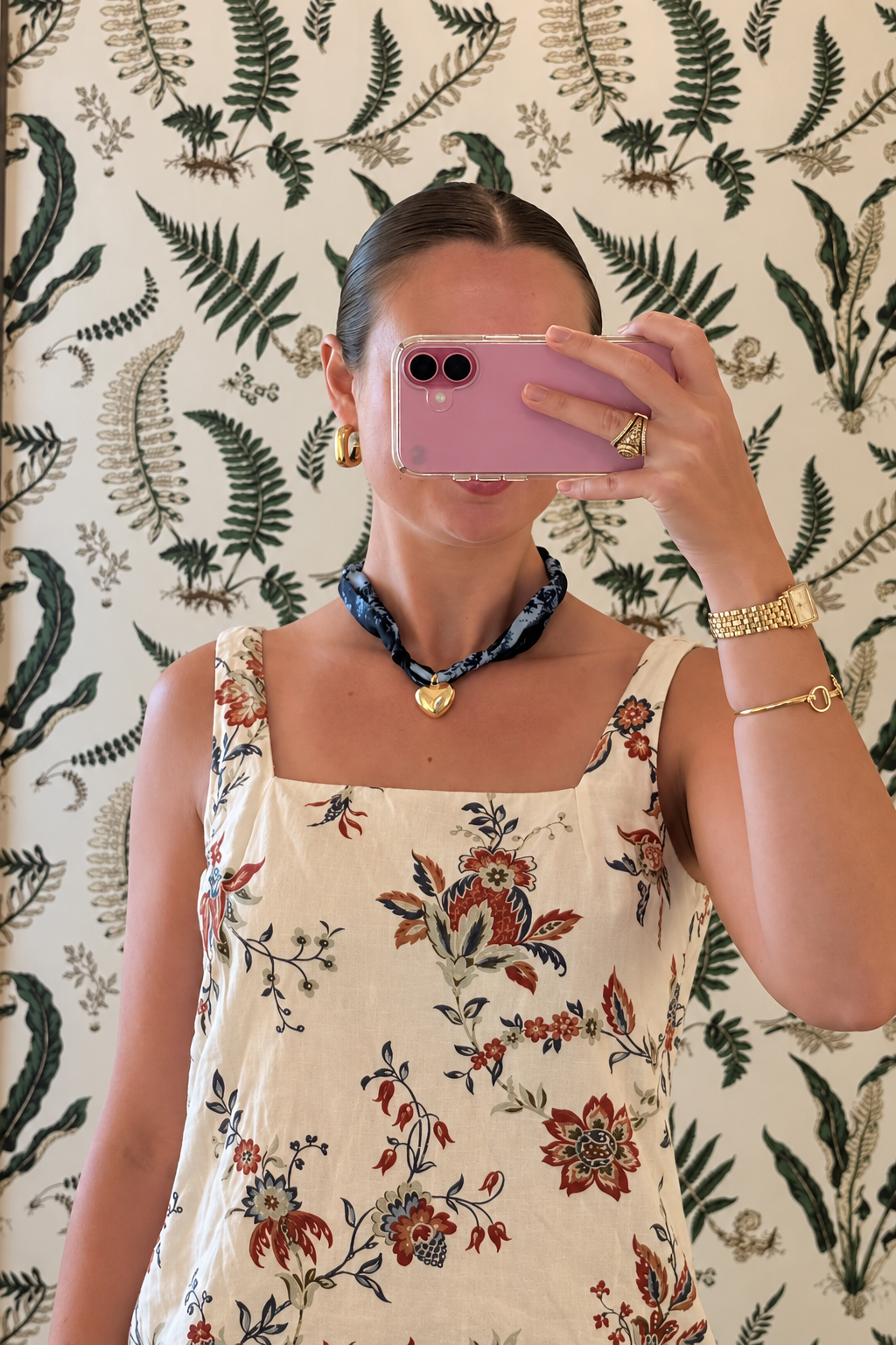 Woman wearing The Andrea silk scarf necklace with a floral print white dress, holding phone, with botanical wall paper in the background